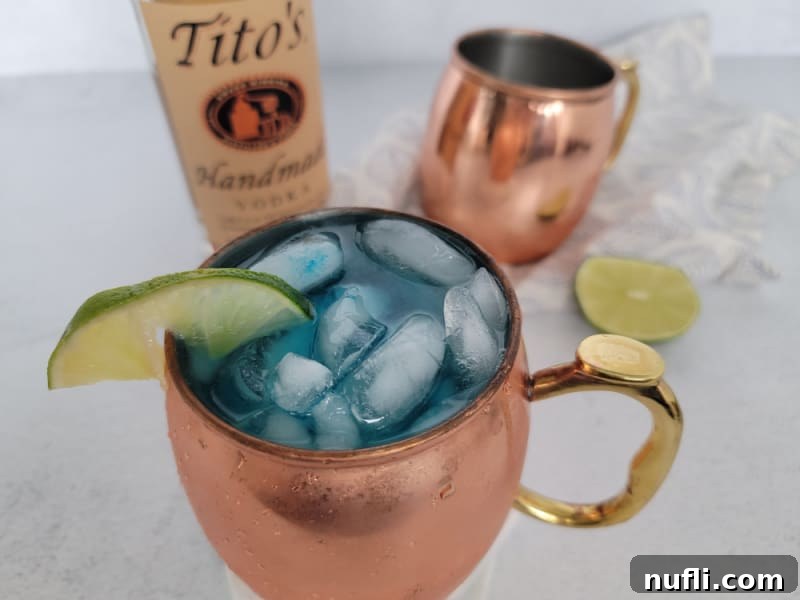 Blue mule cocktail in a copper mug next to a bottle of Tito's and another copper mule mug