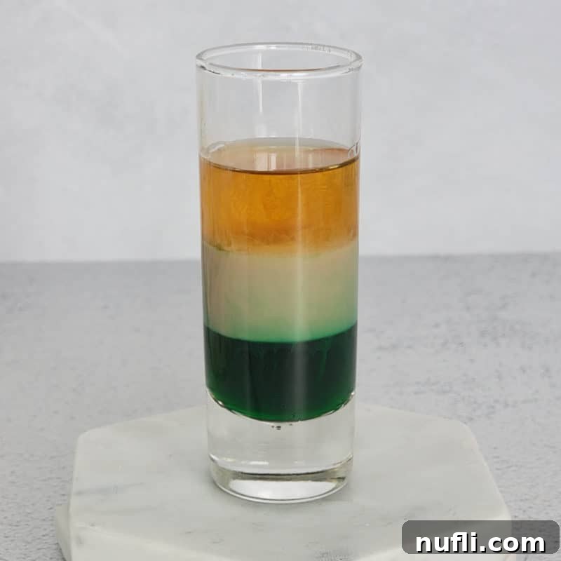 St Patrick's Day Sips & Shots 22 A perfectly layered Irish Flag shot, displaying the distinct green, white, and orange colors, served in a clear glass shot glass on a elegant marble coaster.