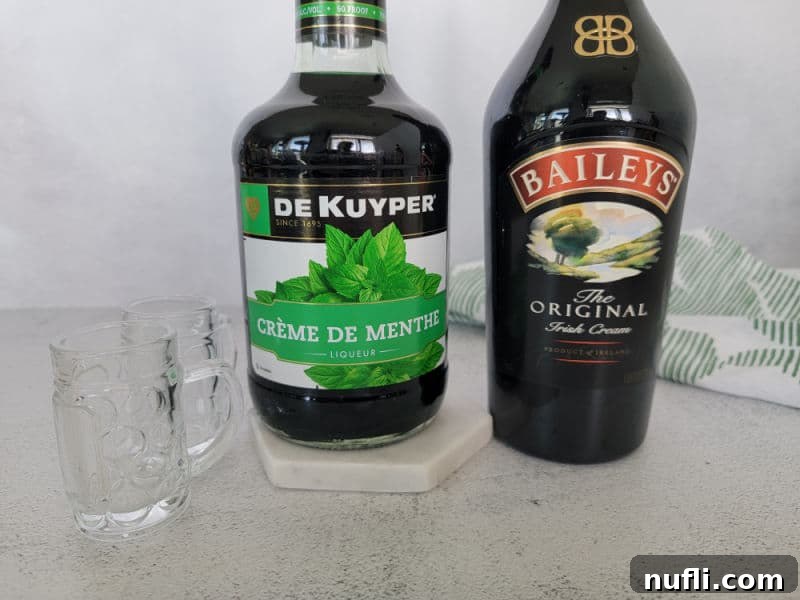 Ingredients for Leprechaun Beer Shot: Green Creme De Menthe and Baileys Irish Cream bottles sit next to two mini beer shot glasses filled with the layered green drink.