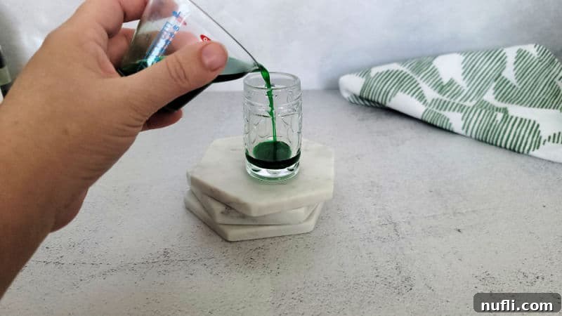 A hand carefully pouring green Creme De Menthe into a mini beer shot glass, placed on white coasters, demonstrating the first step in making a Leprechaun Beer Shot.