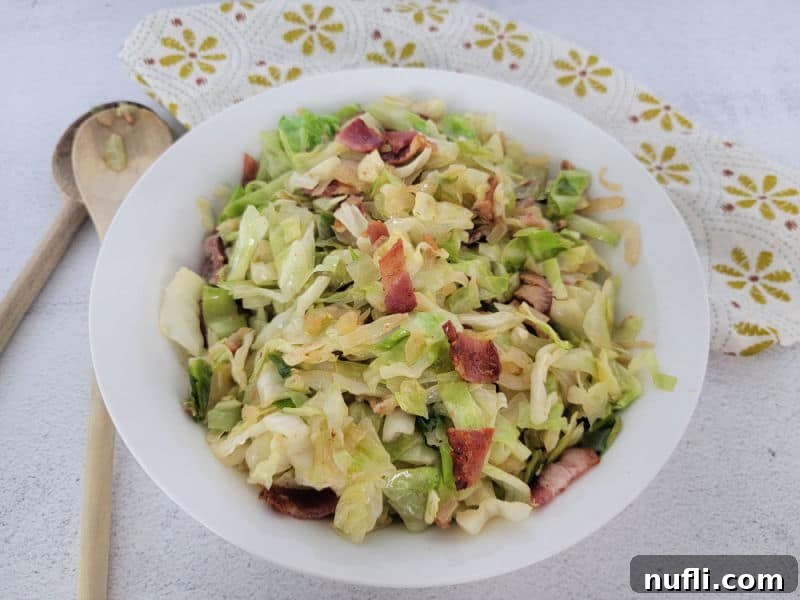 Fried cabbage with bacon in a white bowl next to wooden spoons and a cloth napkin 