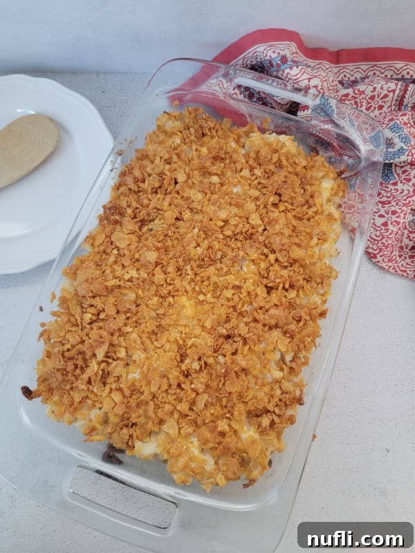 Baked funeral potatoes in a glass baking dish next to a white plate and wooden spoon. 