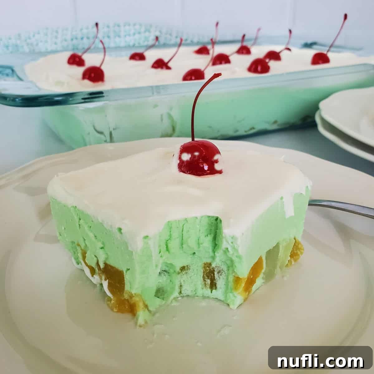 Square of green jello salad garnished with a cherry on a white plate next to a baking dish filled