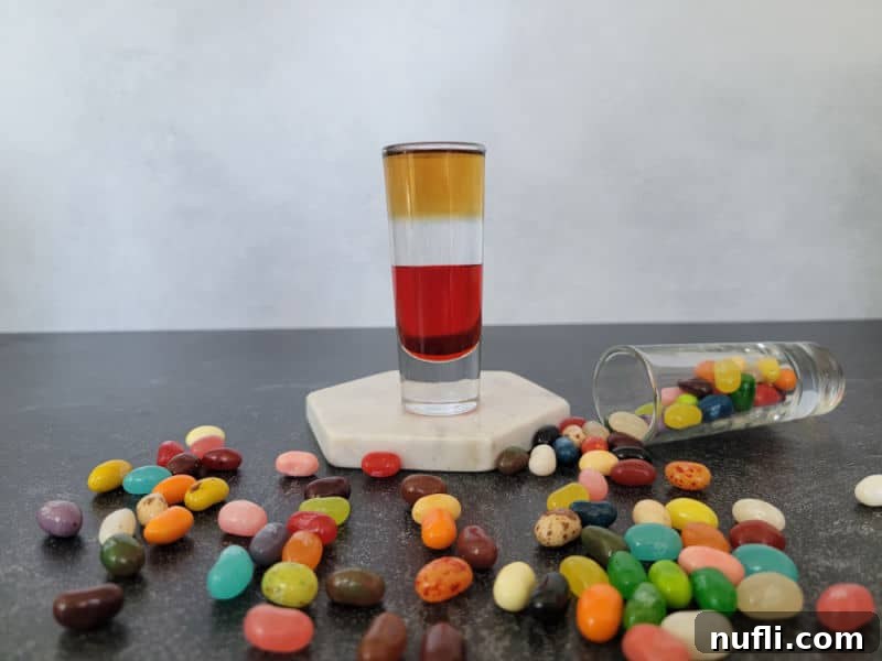 The Art of Grenadine Cocktails 18 A layered Jelly Bean Shot on a white coaster, surrounded by assorted jelly bean candies.