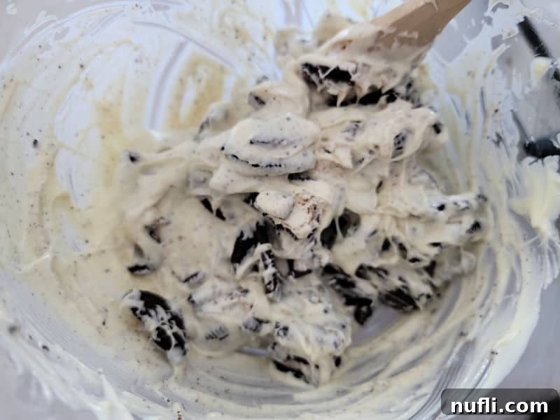 Effortless Two-Ingredient Oreo Bark 6 Oreo cookie pieces being gently folded into melted white chocolate in a glass bowl with a wooden spoon.