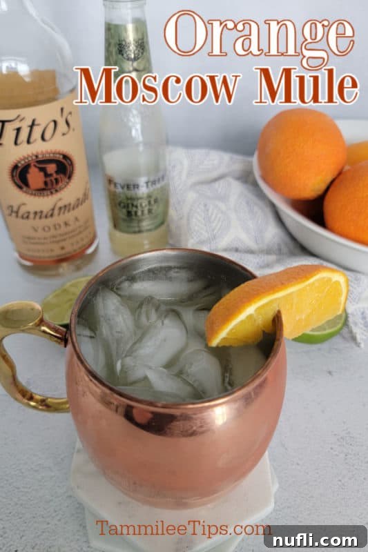 A copper mug filled with an Orange Moscow Mule, garnished with a fresh orange slice, sits beside a bottle of vodka, ginger beer, and a bowl of fresh oranges.