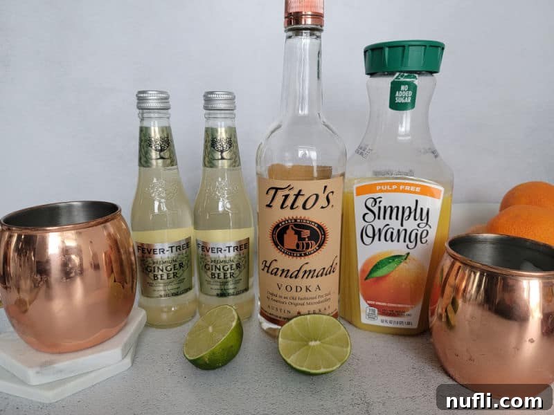 Arrangement of cocktail ingredients including a copper mule mug, bottles of ginger beer and Tito's vodka, a carton of orange juice, and two fresh oranges on a kitchen counter.