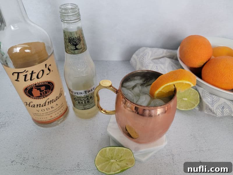 A chilled copper mug with an Orange Moscow Mule and orange slice garnish sits next to bottles of Tito's vodka and ginger beer, with a bowl of oranges in the background.