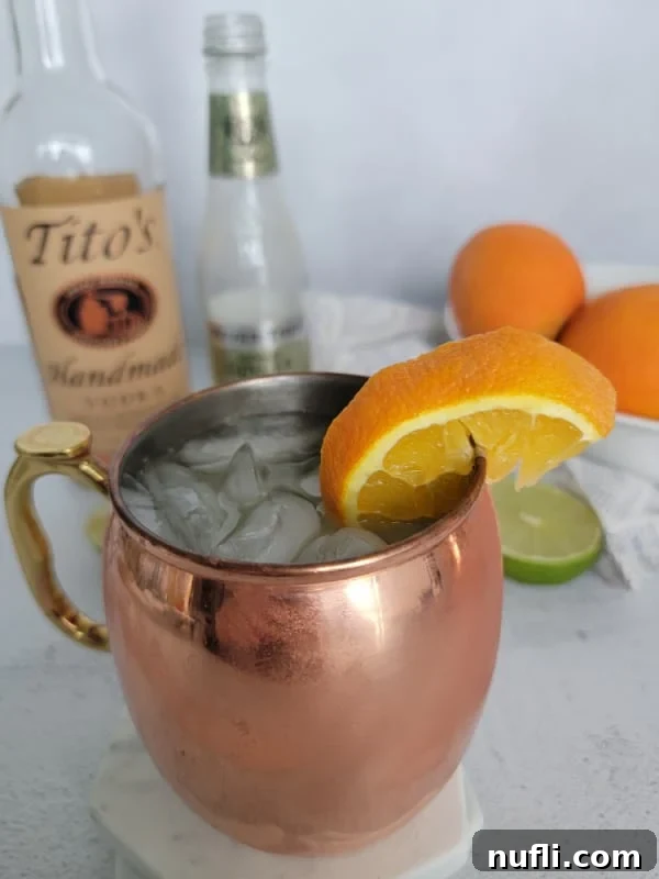 A close-up of an Orange Moscow Mule in a copper mug, garnished with an orange slice. Bottles of vodka and ginger beer are blurred in the background.