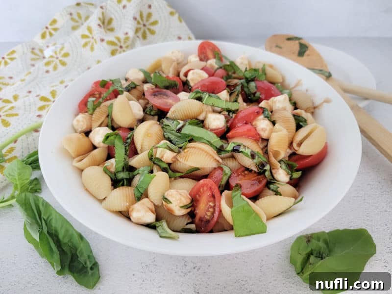 Vibrant Italian Pasta Salad 8 Caprese pasta salad in a white bowl garnished with fresh basil leaves, next to a wooden spoon, emphasizing freshness and simplicity.
