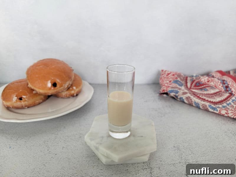 Shot glass half full with creamy liquid next to jelly doughnuts, illustrating the first step of adding RumChata.