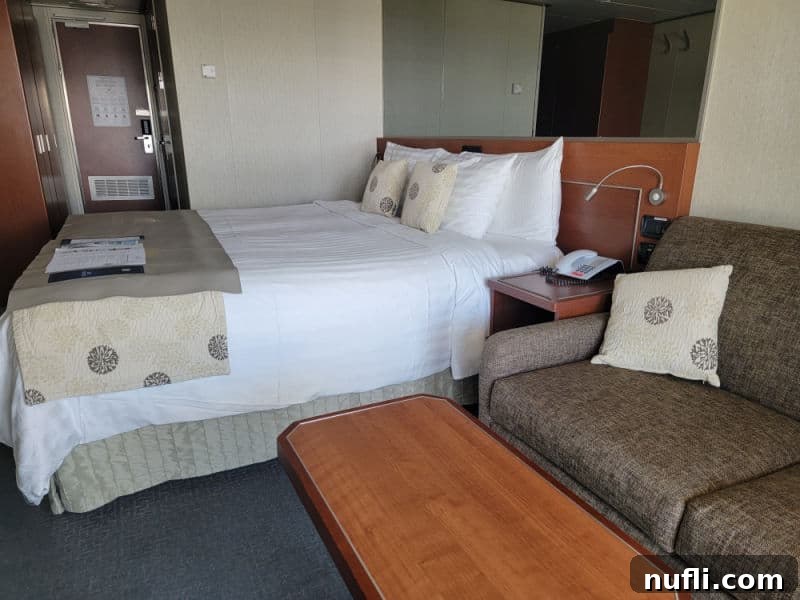 Eurodam stateroom with brown couch and pillow, small table, next to a bed looking towards the door 