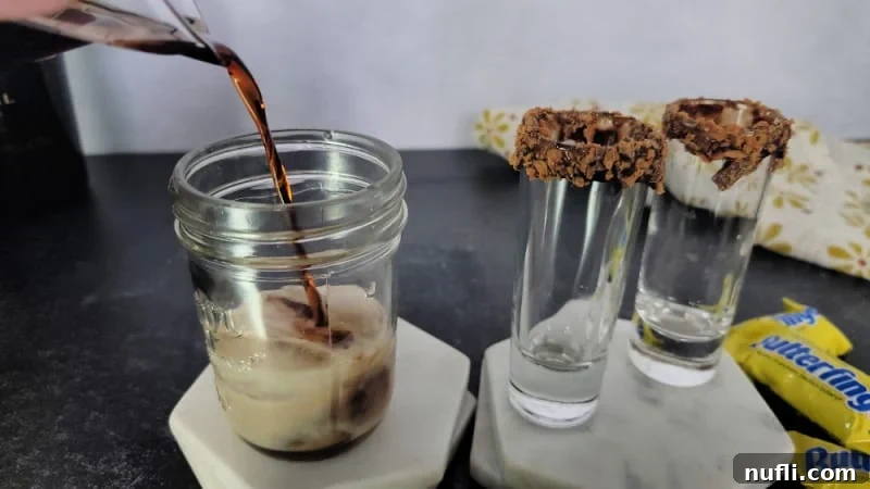 Butterfinger Bliss Shot 5 Brown liquid pouring into a small mason jar on a white coaster next to to shot glasses rimmed in chocolate and Butterfinger pieces