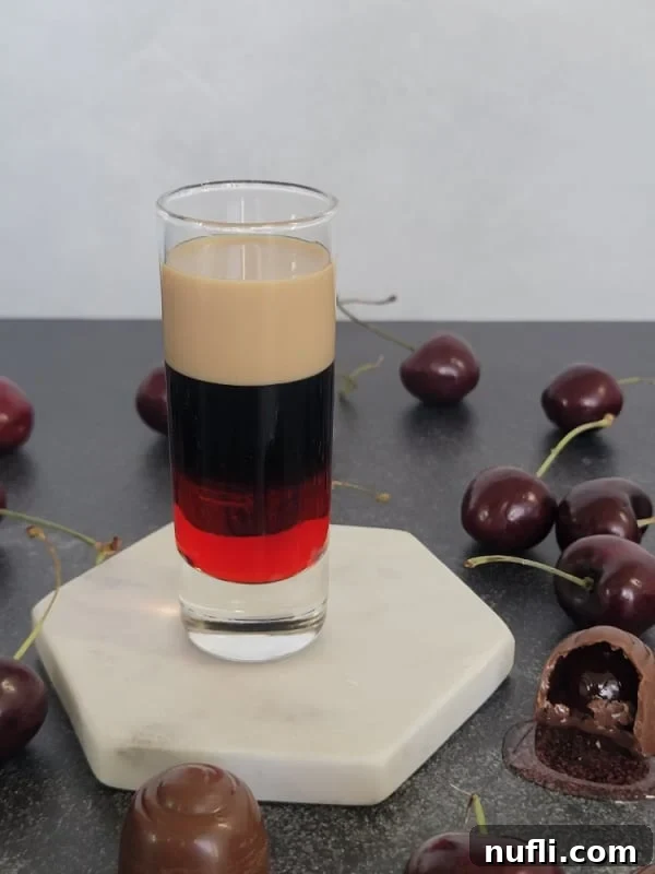 A stunning Chocolate Covered Cherry layered shot, showcasing distinct red, brown, and cream layers in a clear glass, beautifully arranged with chocolate-covered cherry candies.