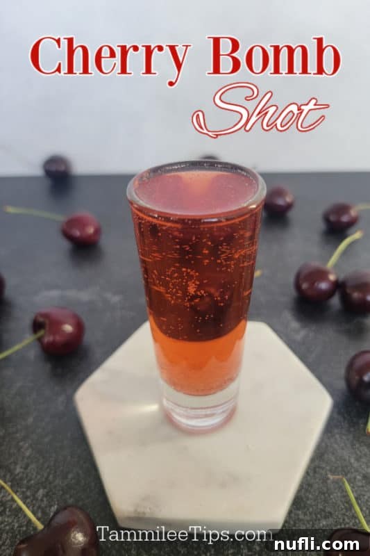 A vibrant red Cherry Bomb Shot in a clear shot glass, garnished with fresh cherries, sitting on a white coaster.