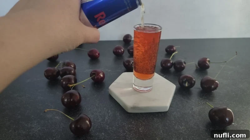 Red Bull being poured into a shot glass already containing red liquid, with fresh cherries scattered around.