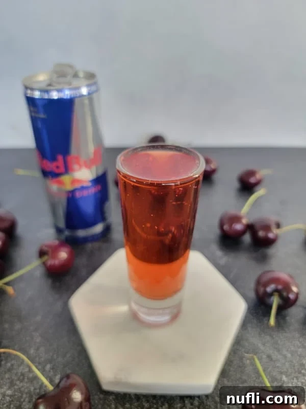 A vibrant red cocktail shot on a white coaster, accompanied by a can of Red Bull and fresh, ripe cherries, highlighting the shot's ingredients and appeal.