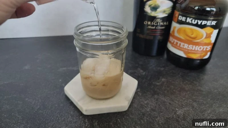 Clear buttershots pouring over Irish Cream in a mason jar glass on a marble coaster
