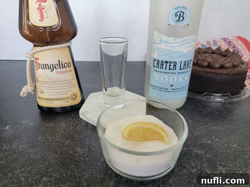 Essential ingredients for a Chocolate Cake Shot: Frangelico, a bottle of vodka, and a lemon wedge in a bowl of sugar, all set beside a chocolate cake.