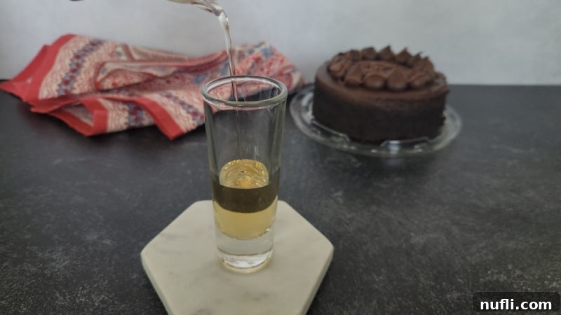 A stream of hazelnut liqueur being poured into a shot glass, with a chocolate cake visible in the background.