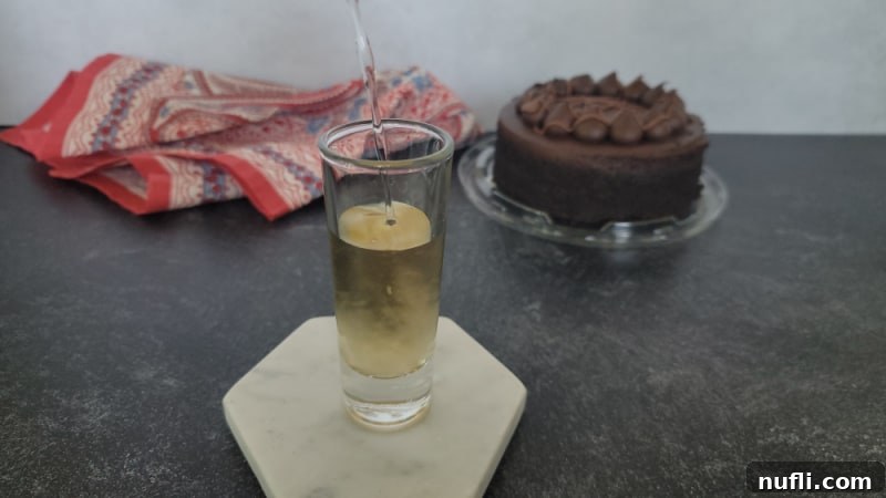 Vodka being carefully poured into a shot glass, completing the liquid components of the Chocolate Cake Shot, next to a chocolate cake.