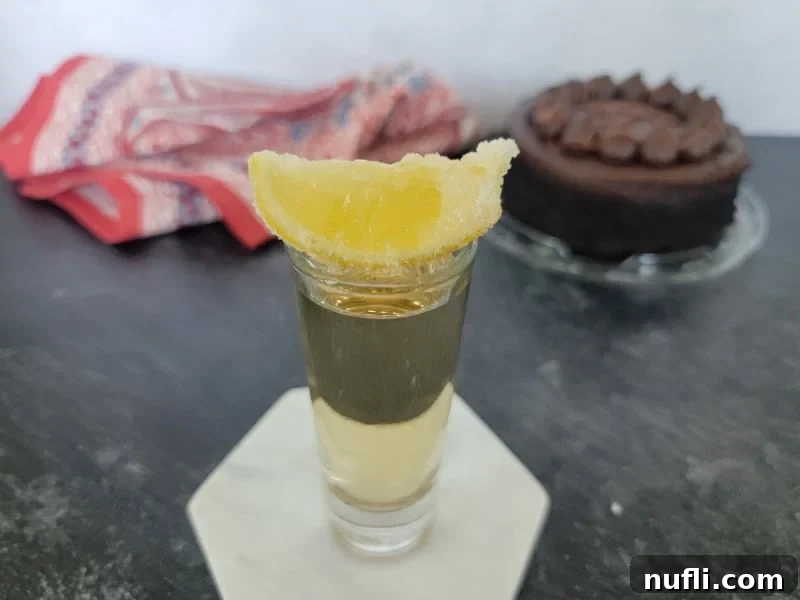 A sugared lemon resting on the rim of a Chocolate Cake Shot glass, positioned elegantly in front of a chocolate cake.
