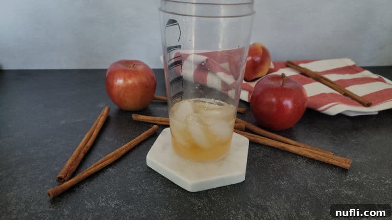 ice and liquid in a cocktail shaker next to cinnamon sticks and apples