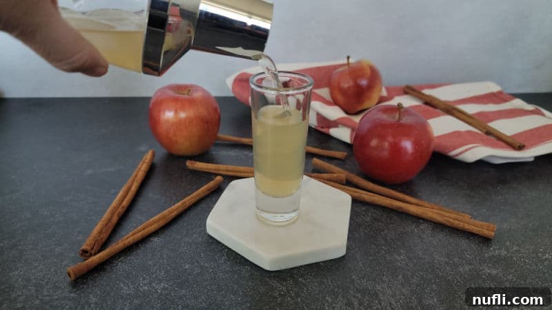Cocktail shaker pouring an apple pie shot into a shot glass on a white coaster next to cinnamon sticks and apples