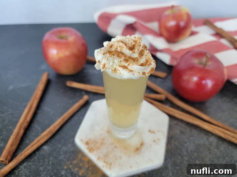 Apple Pie In A Shot Glass 6 Apple pie shot garnished with whipped cream and cinnamon on a white coaster next to cinnamon sticks and red apples