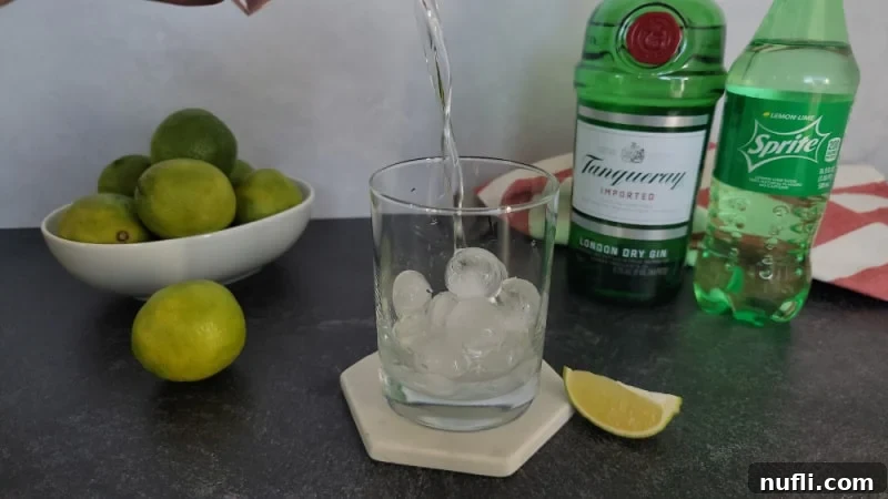 Clear liquid pouring into cocktail glass next to a bowl of limes, bottle of gin, and Sprite