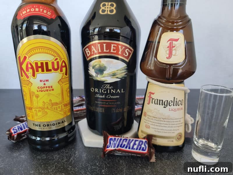 Bottles of Kahlua, Bailey's Irish Cream, and Frangelico lined up next to an empty shot glass, ready for mixing the Snickers shot.