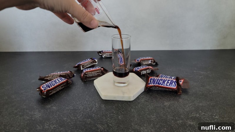 Kahlua pouring into a shot glass, forming the first layer of a Snickers shot, with Snickers Candy Bars in the background.