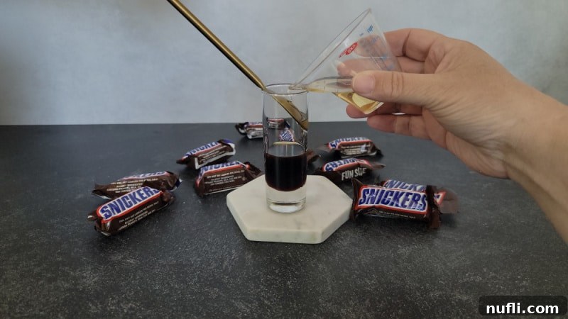 Frangelico pouring over the Kahlua in a shot glass, creating the second distinct layer of the Snickers shot, with Snickers Candy Bars nearby.