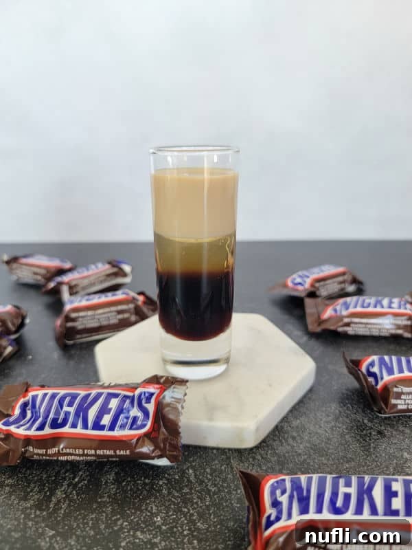 Close-up of a perfectly layered Snickers Shot on a white coaster, with more Snickers Chocolate Bars for visual context.