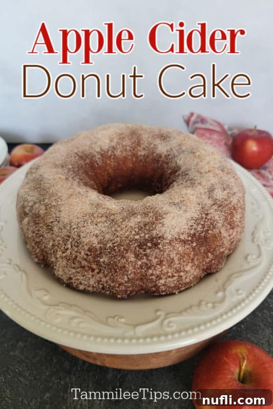 A delicious Apple Cider Donut Cake on a white bundt cake stand, surrounded by fresh apples, ready to be served for a festive fall gathering.