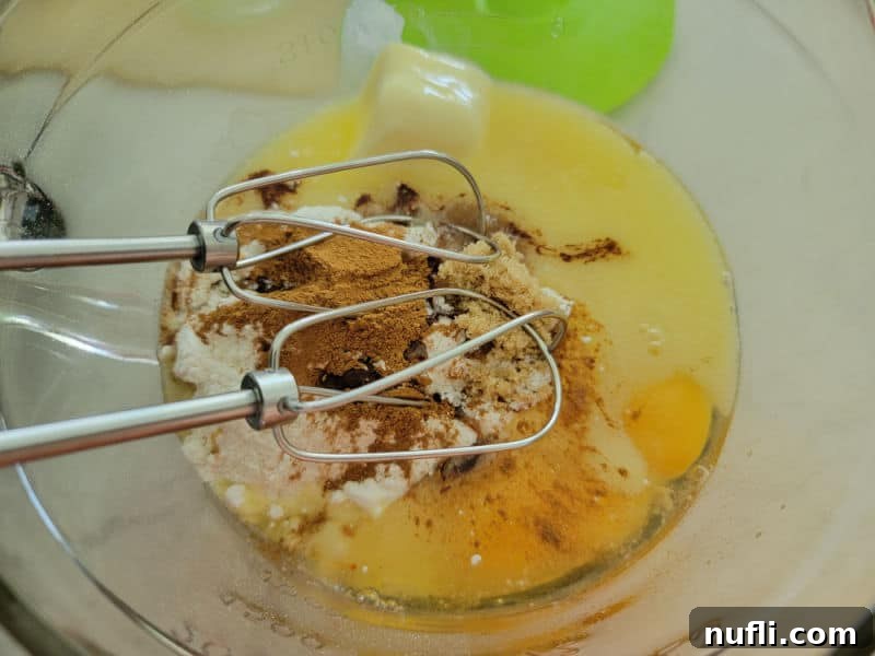 A hand mixer blending cake mix, apple cider, melted butter, eggs, cinnamon, brown sugar, and vanilla extract in a large glass mixing bowl, illustrating the initial step of combining ingredients.