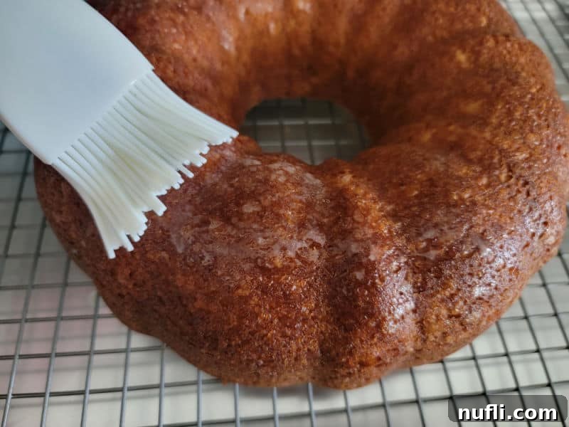 A hand brushing melted butter over the surface of a cooled Apple Cider Donut Cake, preparing it for the cinnamon sugar coating.