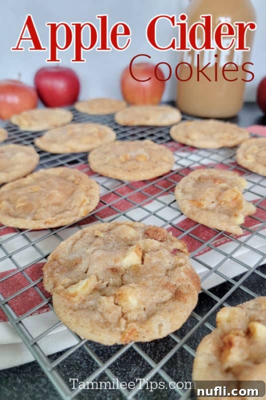 Freshly baked Apple Cider Cookies cooling on a wire rack, with a rustic cloth napkin, a jug of apple cider, and whole apples in the background, creating an inviting autumn scene.