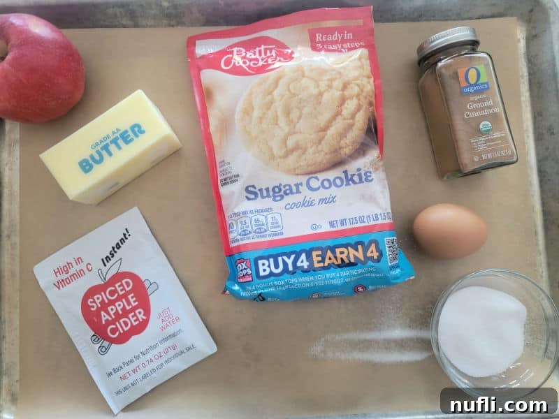 A collection of Apple Cider Cookie ingredients artfully arranged on a parchment-lined baking sheet, including fresh apples, softened butter, a box of sugar cookie mix, dry apple cider mix, cinnamon, an egg, and a bowl of granulated sugar, ready for mixing.