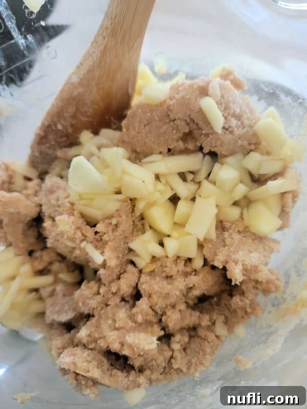 Finely chopped fresh apple pieces being gently stirred into a bowl of dry ingredients, likely a cookie mix, to ensure even distribution for apple cider cookies.