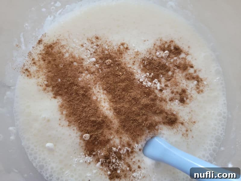 A generous sprinkle of pumpkin pie spice settling onto the light and fluffy angel food cake batter in a glass mixing bowl, ready to be incorporated for rich fall flavor.