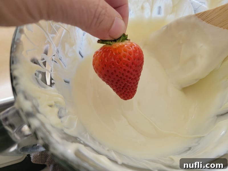 A fresh strawberry being dipped into smooth white chocolate in a clear glass bowl