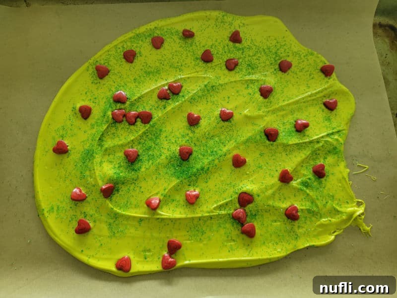 Festive Grinch Bark Made Easy 6 Vibrant Green Candy melt spread out on parchment paper topped with red heart candies and green sanding sugar to make Grinch Bark