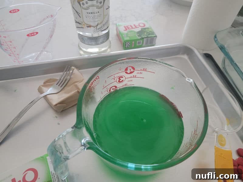 Deliciously Easy Festive Grinch Jello Shots 4 Green jello mixture in a large measuring bowl on a baking sheet, with vodka and a whisk nearby, preparing Grinch Jello Shots