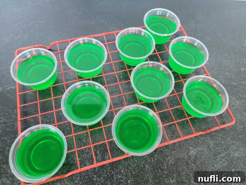 Deliciously Easy Festive Grinch Jello Shots 5 Rows of bright green jello shots in plastic cups, arranged on a red cooling rack, ready for garnishing