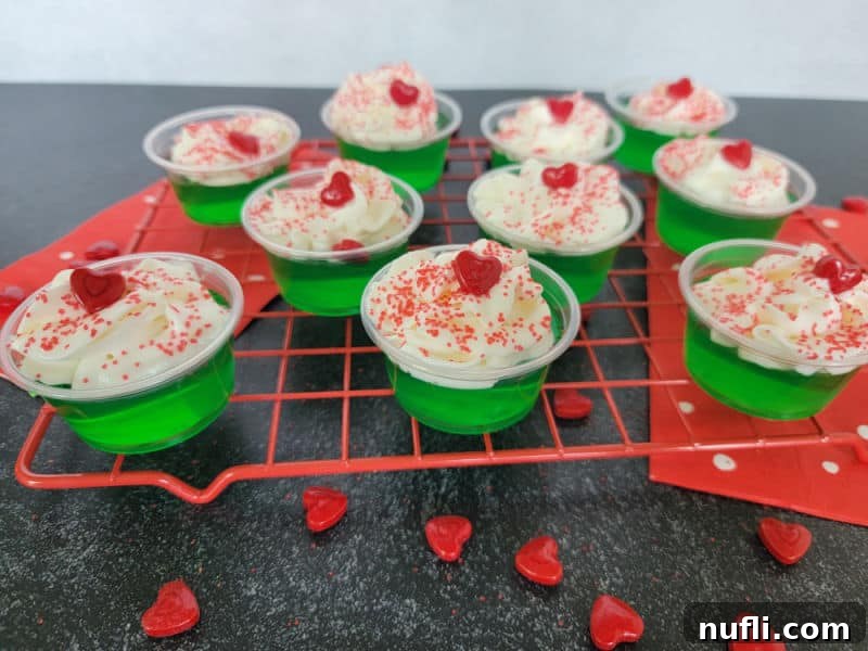 Deliciously Easy Festive Grinch Jello Shots 6 Finished Grinch Jello Shots, beautifully garnished with whipped cream, festive red sprinkles, and a small red candy heart, resting on a red drying rack
