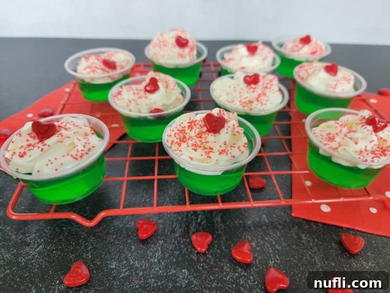 Deliciously Easy Festive Grinch Jello Shots 7 Close-up of green Grinch Jell-O Shots, garnished with white whipped cream, vibrant red heart candies, and colorful sprinkles, creating a festive holiday look