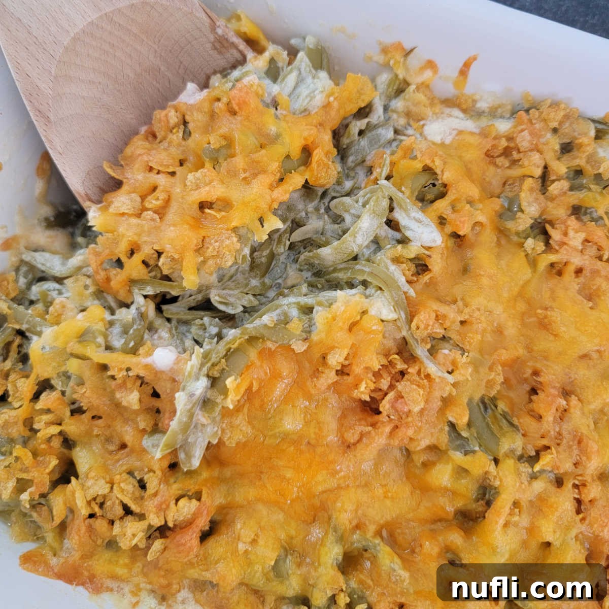 A wooden spoon scooping a generous portion of cheesy green beans from a white casserole dish, showcasing its creamy texture.