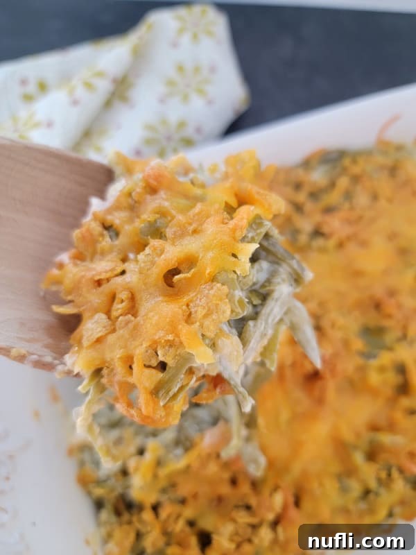 A close-up of cheesy green beans on a wooden spoon, held above the casserole dish, showing the crispy topping and creamy interior.