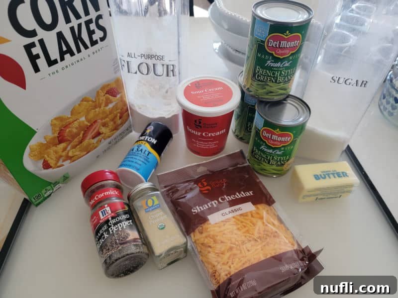 Ingredients for Cheesy Green Beans including cornflakes, flour, sugar, sour cream, green beans, butter, cheddar cheese, salt, pepper, and garlic powder arranged on a surface.